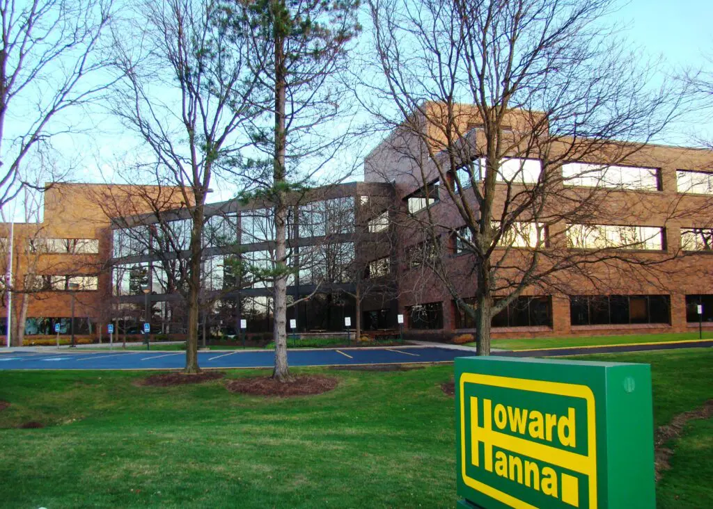 A brick building behind leafless trees and a lawn with a Howard Hanna sign.